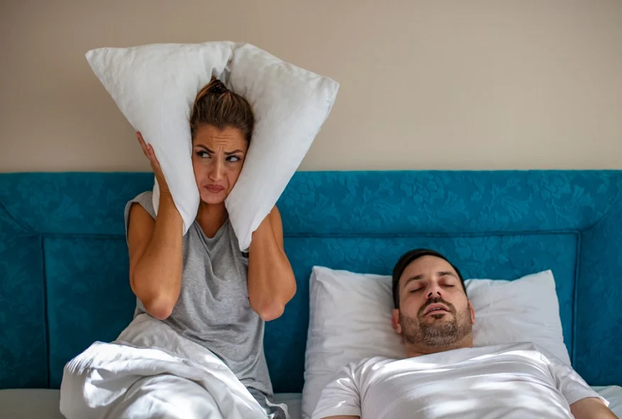 Como a Aliança do Sono pode melhorar o sono do casal? Mulher cobre os ouvidos com o travesseiro enquanto o parceiro ronca. Psicólogo Elídio Almeida – Terapia de Casal Salvador.